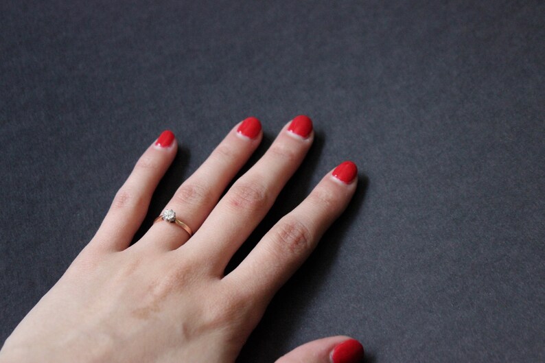 May include: A hand with red nail polish and a gold ring with a single diamond on the ring finger.