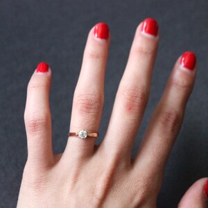 May include: A close-up of a hand with red nail polish wearing a simple gold band with a single round diamond.