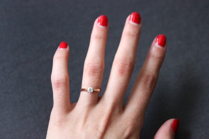 May include: A close-up of a hand with red nail polish wearing a simple gold band with a single round diamond.
