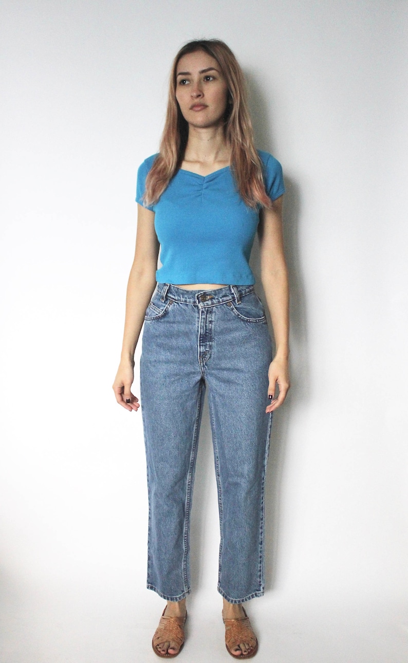 May include: A woman is wearing a light blue cropped t-shirt and blue denim jeans. She is standing on a white background.