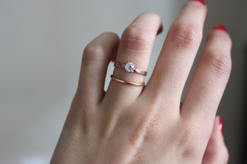 May include: A close-up of a hand wearing two gold rings. The top ring has a single, round, clear gemstone. The bottom ring is a simple band.