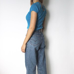 May include: A woman wearing a light blue cropped t-shirt with a criss-cross back detail and blue denim jeans.