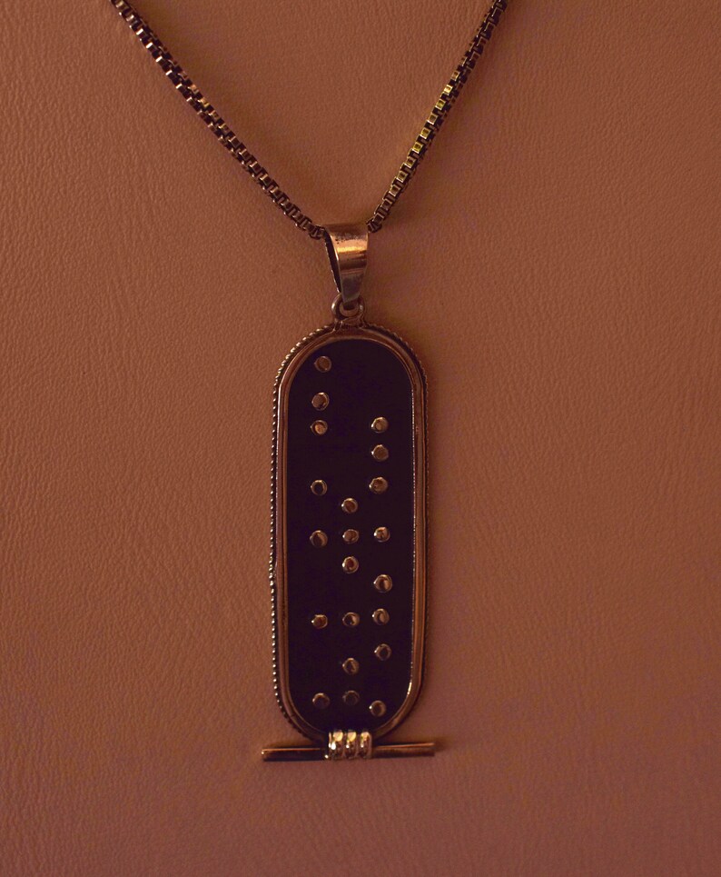 May include: A gold chain necklace with a black pendant featuring a pattern of gold dots. The pendant is rectangular with rounded edges and has a small gold bar at the bottom.