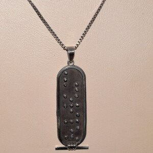 May include: A silver chain necklace with a silver pendant. The pendant is oval shaped and has a pattern of small circles on it. The pendant has a small bar at the bottom.