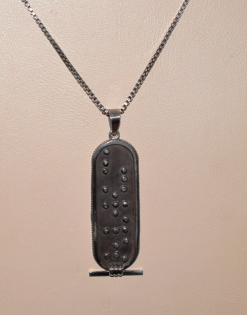 May include: A silver chain necklace with a silver pendant. The pendant is oval shaped and has a pattern of small circles on it. The pendant has a small bar at the bottom.