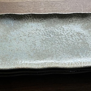 Op de afbeelding: Een stapel van drie rechthoekige keramische borden. De borden hebben een lichtgrijze, getextureerde oppervlakte met een licht onregelmatige, golvende rand. De borden zijn op elkaar gestapeld.