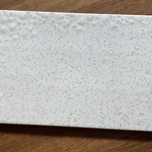 May include: Rectangular white serving board with a speckled pattern. The board has rounded corners and a textured surface. The speckled pattern is a mix of light and dark gray dots, creating a subtle contrast. The board is made of a solid material.