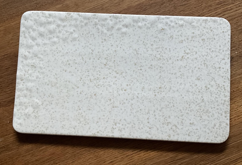 May include: Rectangular white serving board with a speckled pattern. The board has rounded corners and a textured surface. The speckled pattern is a mix of light and dark gray dots, creating a subtle contrast. The board is made of a solid material.