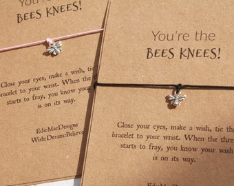 May include: Close-up of two wish bracelets on brown cards. One bracelet has a pink string and a flower charm, the other has a black string and a bee charm. The cards read "You're the Bees Knees!" and include instructions.