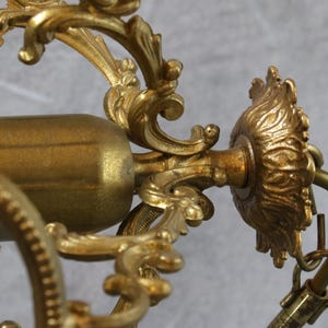 May include: A gold-toned metal chandelier component featuring intricate scrollwork and acanthus leaf detailing. The structure includes a central cylindrical light socket housing and a decorative circular mounting plate with a curved metal hook attachment.