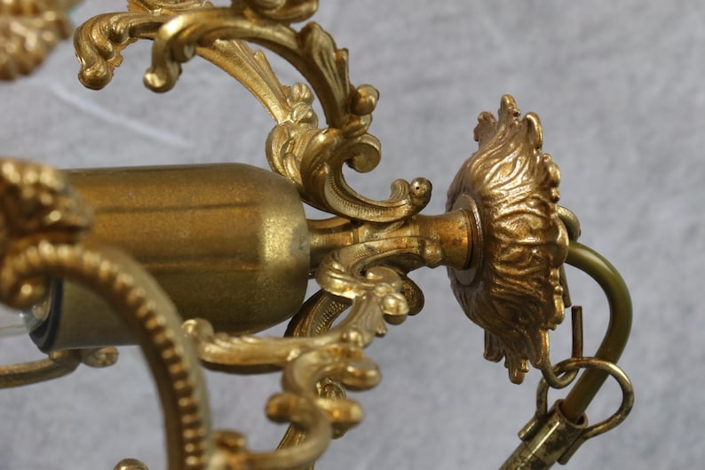 May include: A gold-toned metal chandelier component featuring intricate scrollwork and acanthus leaf detailing. The structure includes a central cylindrical light socket housing and a decorative circular mounting plate with a curved metal hook attachment.