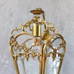 May include: A gold-toned metal lantern pendant light featuring an ornate, scrollwork frame with intricate floral detailing. The fixture includes clear glass panels, a central light bulb socket, and a decorative top canopy with a matching metal chain.