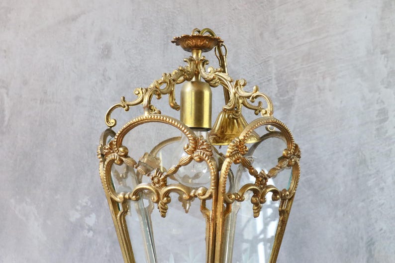 May include: A gold-toned metal lantern pendant light featuring an ornate, scrollwork frame with intricate floral detailing. The fixture includes clear glass panels, a central light bulb socket, and a decorative top canopy with a matching metal chain.