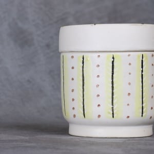 Op de afbeelding: Witte keramische pot met deksel, met een geel en zwart gestreept patroon met kleine rode stippen.