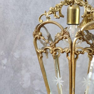 May include: A brass lantern pendant light featuring an ornate, scrolled metal frame with clear glass panels. Each glass panel is etched with a white star-shaped floral design. The fixture includes a central brass socket for a single light bulb.