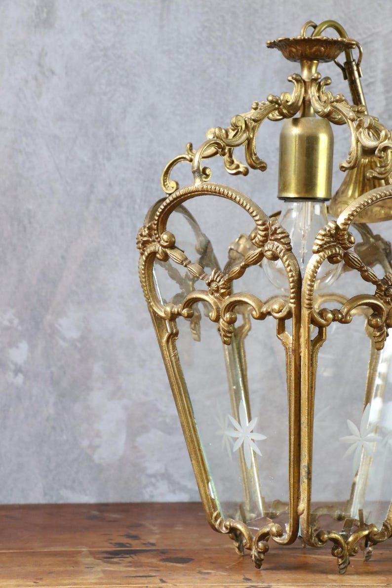May include: A brass lantern pendant light featuring an ornate, scrolled metal frame with clear glass panels. Each glass panel is etched with a white star-shaped floral design. The fixture includes a central brass socket for a single light bulb.