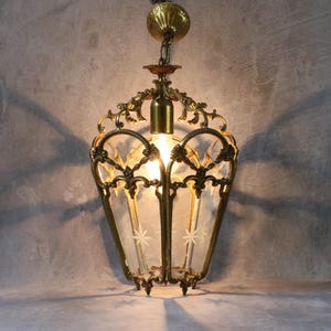 May include: A hanging pendant light fixture featuring an ornate brass frame with intricate scrollwork and clear glass panels. Each glass panel is etched with a small star design. The fixture includes a brass ceiling canopy and a central light bulb.