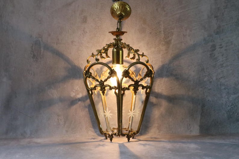 May include: A hanging pendant light fixture featuring an ornate brass frame with intricate scrollwork and clear glass panels. Each glass panel is etched with a small star design. The fixture includes a brass ceiling canopy and a central light bulb.