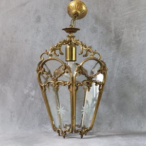 May include: A hanging pendant light fixture featuring a brass frame with ornate scrollwork and clear glass panels. The glass panels are etched with star-shaped designs. The fixture includes a brass ceiling canopy and a central light socket.