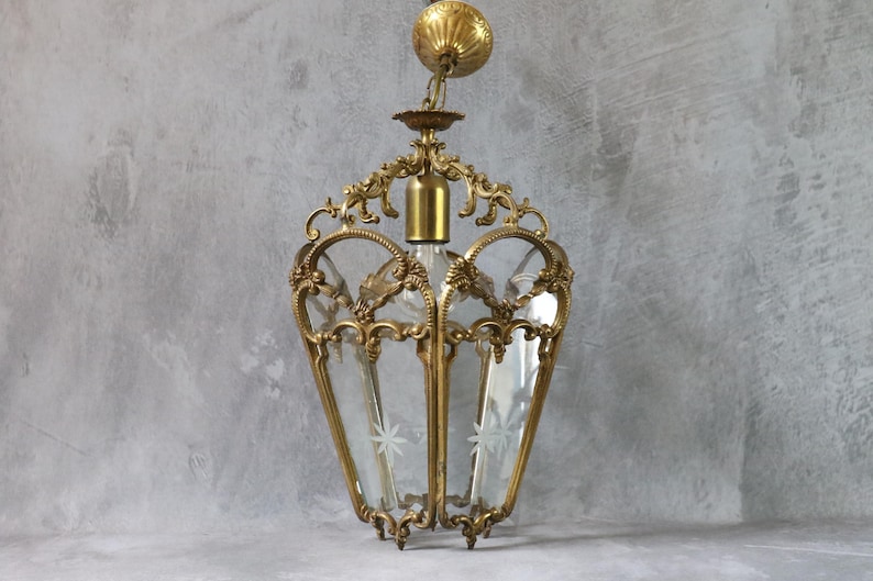 May include: A hanging pendant light fixture featuring a brass frame with ornate scrollwork and clear glass panels. The glass panels are etched with star-shaped designs. The fixture includes a brass ceiling canopy and a central light socket.