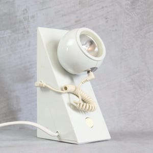 May include: A white desk lamp with a triangular base and a spherical, adjustable light head. The lamp features a coiled cord connecting the head to the base, a circular power button on the front, and a straight white power cable extending outward.