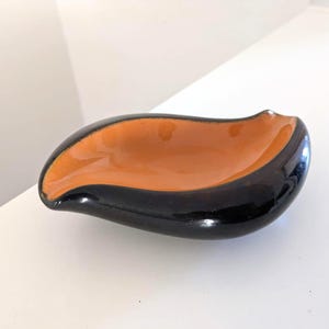 May include: A small, asymmetrical ceramic dish with a smooth, glossy finish. The interior of the bowl is a vibrant orange color, while the exterior and the wide, curved rim are a deep, solid black. The shape is organic, resembling a leaf or petal.