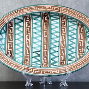 Pode incluir: Uma cesta oval de cerâmica com um design de treliça vazada. A peça apresenta um esmalte verde-azulado com listras verticais marrons decoradas com um padrão de grega branco. A borda também é adornada com um motivo de grega branco correspondente.