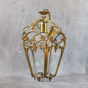 May include: A hanging lantern light fixture featuring an ornate brass frame with intricate scrollwork and decorative metal accents. The lantern has clear glass panels, each etched with a delicate star pattern, and a central brass socket for a light bulb.