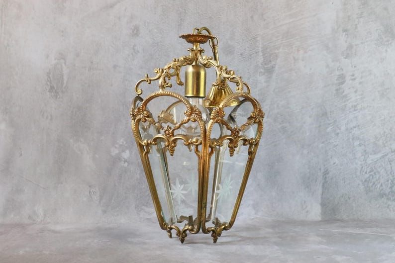 May include: A hanging lantern light fixture featuring an ornate brass frame with intricate scrollwork and decorative metal accents. The lantern has clear glass panels, each etched with a delicate star pattern, and a central brass socket for a light bulb.