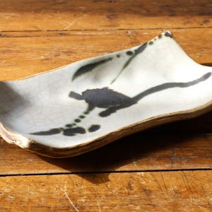 May include: A rectangular ceramic tray with curved, raised edges featuring a light off-white glaze with a visible crackle pattern. The surface is decorated with a dark, abstract ink-like brushstroke design that stretches across the center.