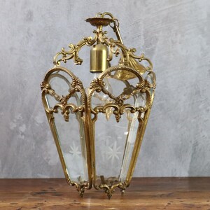 May include: A brass lantern pendant light featuring an ornate metal frame with intricate scrollwork and clear glass panels. The glass panels are etched with star-shaped patterns. A central brass socket is positioned inside the lantern structure.
