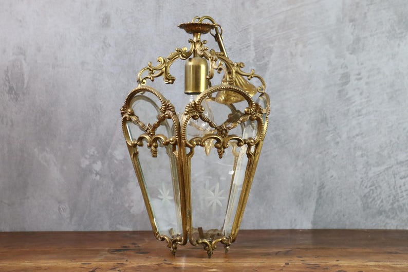 May include: A brass lantern pendant light featuring an ornate metal frame with intricate scrollwork and clear glass panels. The glass panels are etched with star-shaped patterns. A central brass socket is positioned inside the lantern structure.