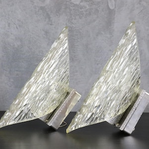 May include: A pair of wall sconces featuring triangular, clear glass shades with a textured, rippled surface. Each shade is mounted on a rectangular, metallic silver base with a decorative, embossed pattern along the edges of the fixture.