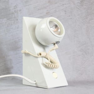 Puede incluir: Una lámpara de escritorio blanca de estilo retro con una lámpara esférica. La lámpara tiene una base triangular y un cable en espiral. La lámpara está inclinada y enchufada a una toma de corriente.