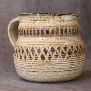 Peut inclure: Un pichet en céramique avec un émail moucheté crème et brun. Le corps présente des bandes horizontales de texture incisée, dont un motif entrecroisé proéminent au centre et un motif de lignes verticales près du bord supérieur.