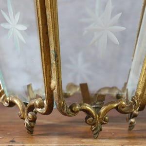 May include: A gold-toned metal lantern frame with ornate, curved base detailing and clear glass panels. Each glass pane features a frosted, etched starburst pattern. The structure is hexagonal in shape with visible metal joints and edges.