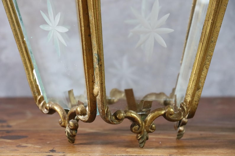 May include: A gold-toned metal lantern frame with ornate, curved base detailing and clear glass panels. Each glass pane features a frosted, etched starburst pattern. The structure is hexagonal in shape with visible metal joints and edges.
