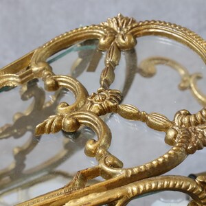 May include: A gold-toned metal frame with intricate scrollwork and floral detailing surrounding a clear glass panel. The metal structure features curved lines and ornate decorative elements that form a border around the transparent glass surface.