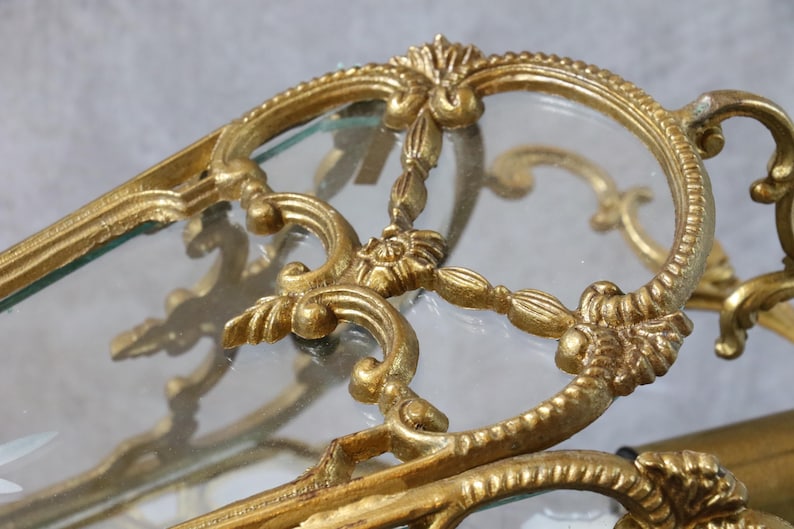 May include: A gold-toned metal frame with intricate scrollwork and floral detailing surrounding a clear glass panel. The metal structure features curved lines and ornate decorative elements that form a border around the transparent glass surface.