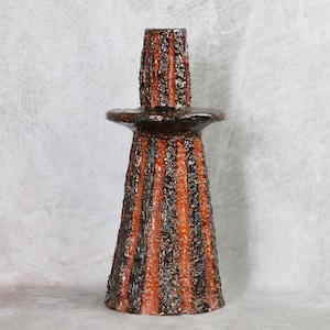 May include: A textured, ceramic candle holder with a dark brown and orange striped design. The candle holder has a wide base that tapers to a narrower top, with a small, round platform in the middle. The surface has a rough, uneven texture.