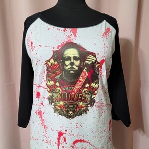 Könnte beinhalten: Ein weißes und schwarzes Baseball-Shirt mit rotem Spritzmuster. Das Shirt zeigt eine Grafik eines Mannes mit einem Messer und den Text "Willkommen zu Halloween".