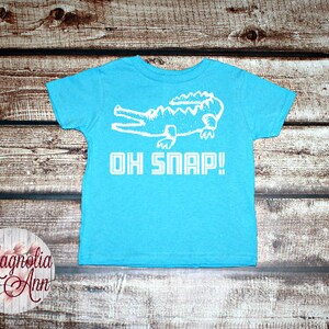 May include: A turquoise blue t-shirt with a white alligator graphic and the text "OH SNAP!" printed on the front.