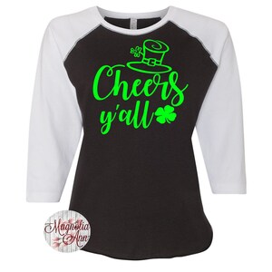以下が含まれることがあります： 黒と白のベースボールスタイルのTシャツで、緑色のテキストに「Cheers y'all」と、緑色のクローバーとレプリコーンの帽子が描かれています。