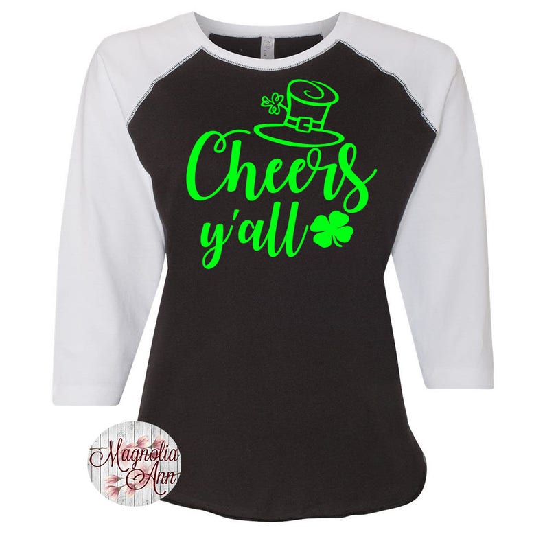 以下が含まれることがあります： 黒と白のベースボールスタイルのTシャツで、緑色のテキストに「Cheers y'all」と、緑色のクローバーとレプリコーンの帽子が描かれています。