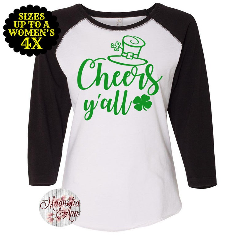 以下が含まれることがあります： 白と黒のベースボールTシャツに、緑色の文字で「Cheers y'all」と書いてあり、緑色のレプリコンの帽子にクローバーが飾られています。