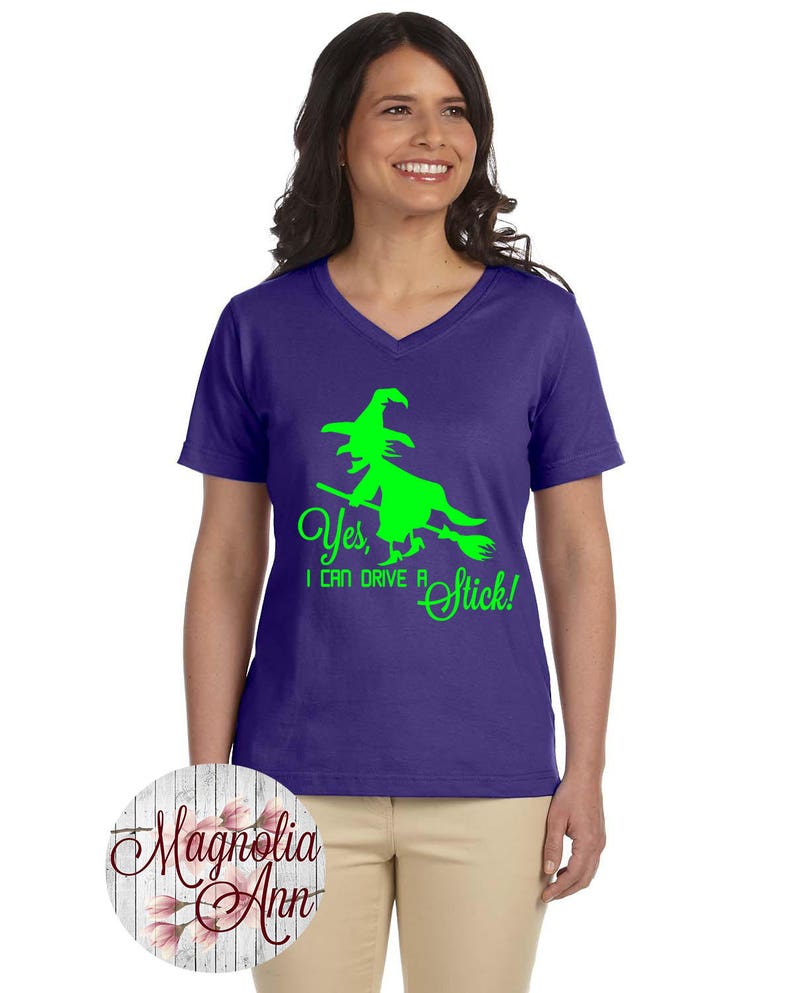 May include: Purple V-neck t-shirt with a green witch silhouette riding a broomstick and the text "Yes, I can drive a stick!"