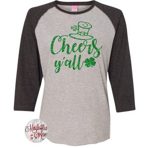 以下が含まれることがあります： グレーとブラックのベースボールTシャツに、緑色のグリッターで「Cheers y'all」と書いてあり、緑色のグリッターのクローバーと緑色のグリッターのレプリコーンの帽子が付いています。