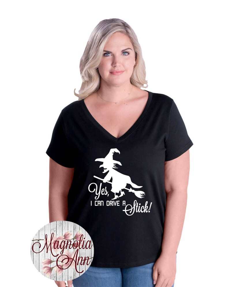May include: Black v-neck t-shirt with a white graphic of a witch flying on a broomstick. The text "Yes, I can drive a stick!" is printed below the graphic.