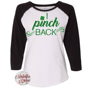 May include: A white and black baseball tee shirt with green glitter text that reads "I pinch back" with a shamrock.