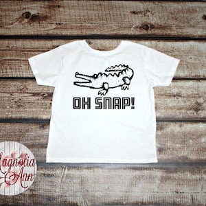 May include: White t-shirt with a black and white illustration of an alligator and the text "Oh Snap!"